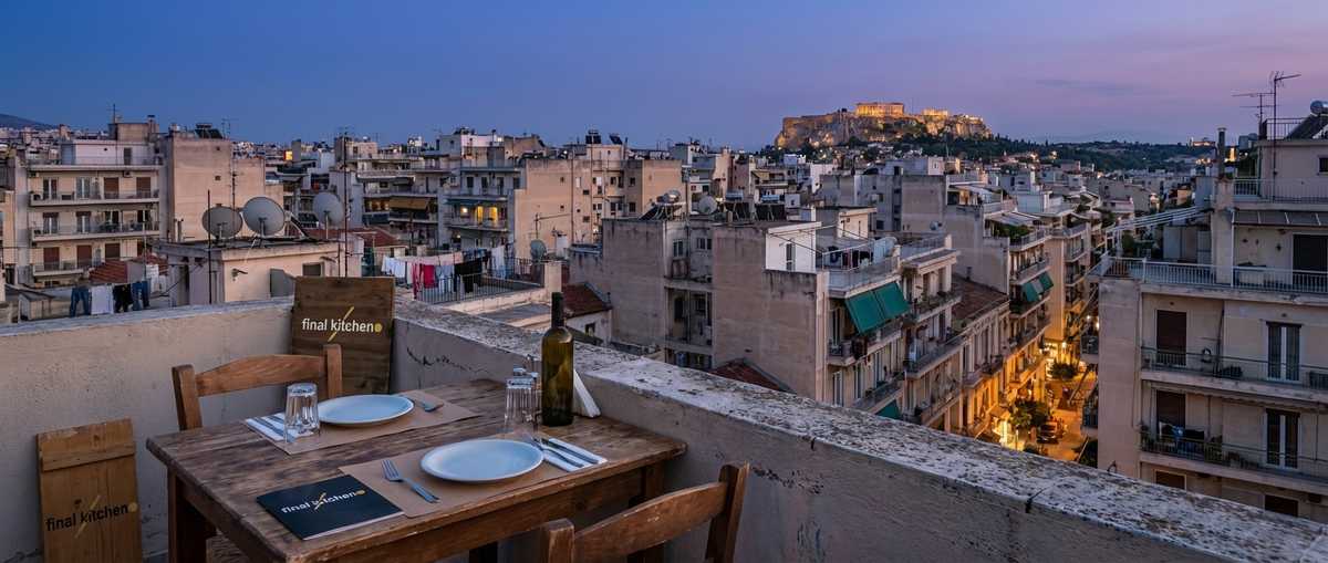 Athens rooftop at dusk — apartments, satellite dishes, the Acropolis in the distance, a table set for two